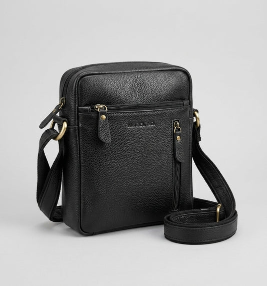 A black genuine leather crossbody bag with a front zippered pocket, brass-tone hardware, and a detachable shoulder strap, shown on a light gray background.