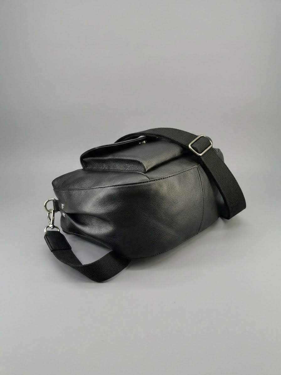 Black leather shoulder bag with a wide adjustable strap and silver buckle, photographed on a neutral gray background.