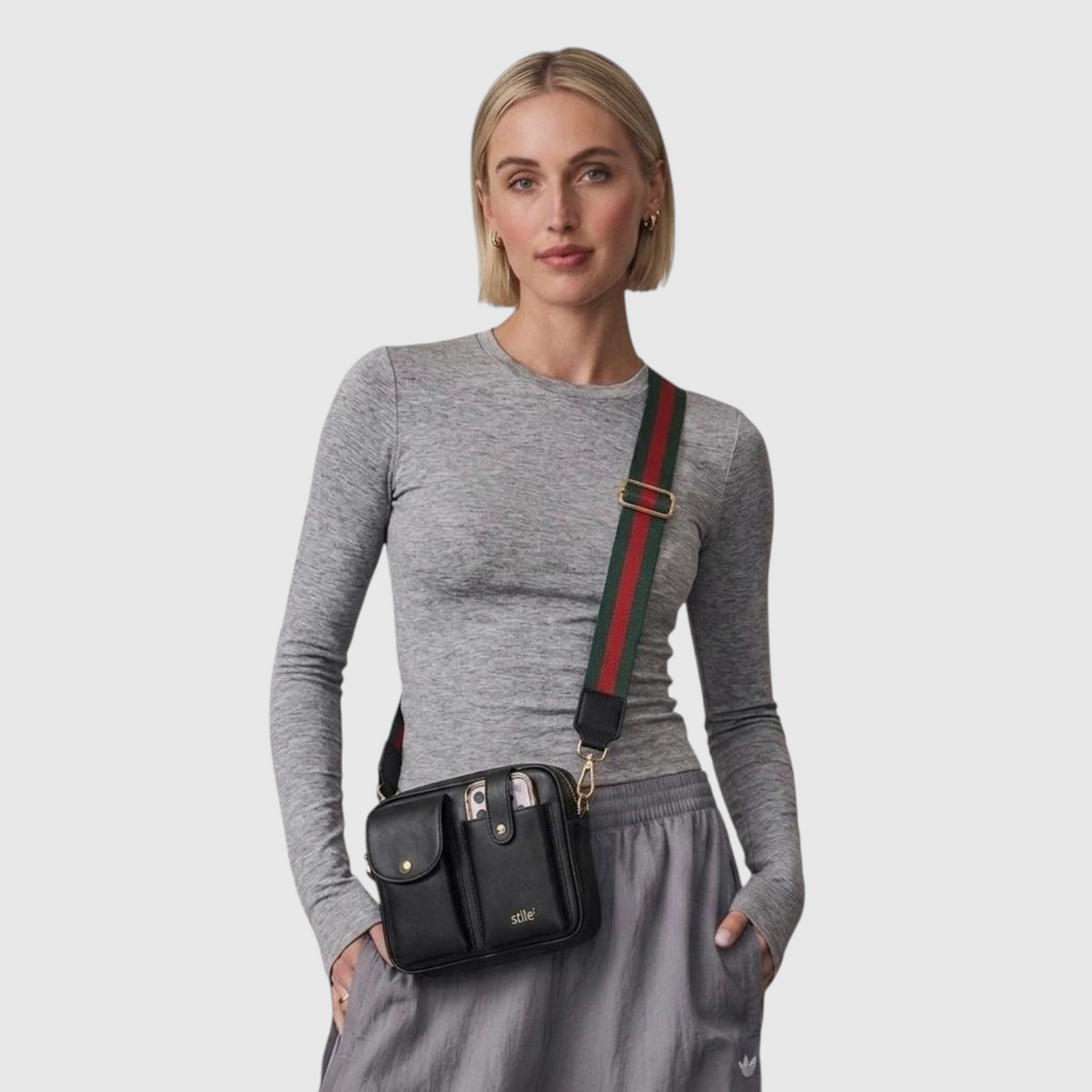A woman wearing a gray long-sleeve top and gray sweatpants models a black crossbody bag with a green and red web stripe strap and gold hardware against a light gray background.