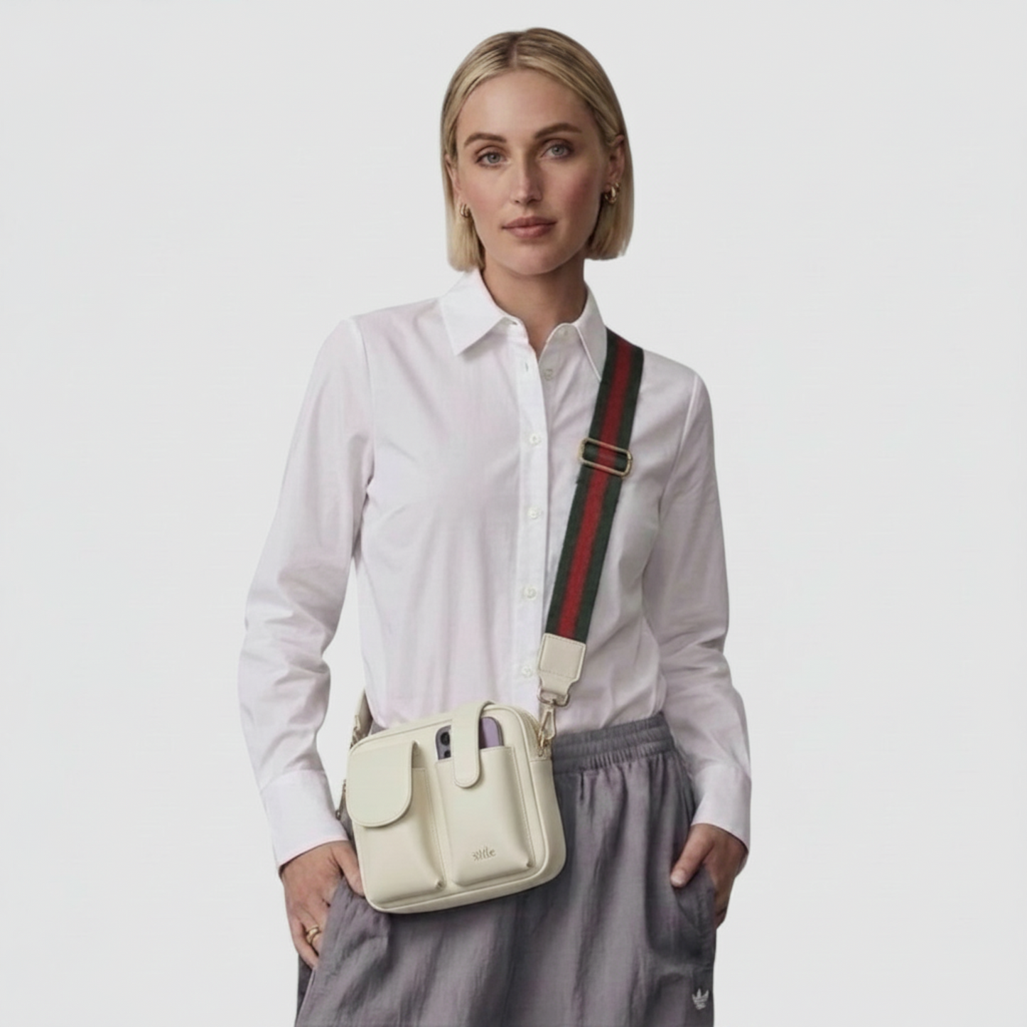 A woman wearing a white button-down shirt and gray trousers models an Ivory-colored crossbody bag with a signature green and red web stripe strap, set against a plain light gray background.